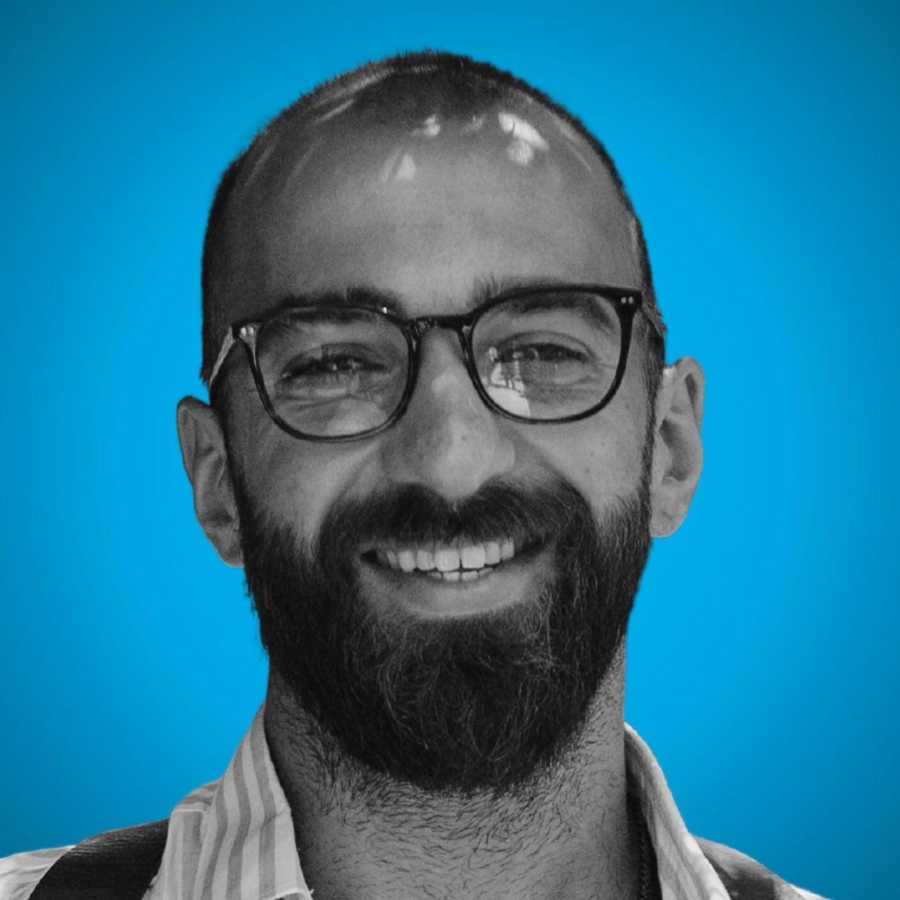 Porträt eines lächelnden Mannes mit Brille und Vollbart auf einem blauen Hintergrund